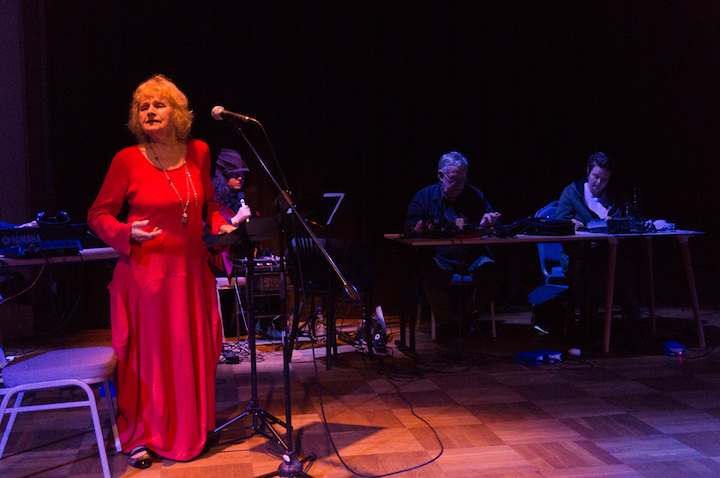 Koncert. W pierwszym planie śpiewaczka Olga Szwajgier w czerwonej sukni, w tle muzycy z instrumentami elektronicznymi