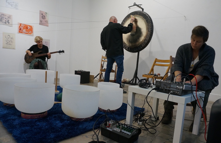 zdjęcie z koncertu. Po lewej gitarzystka, w centrum artysta grający na gongach a po prawej artystka grająca na instrumentach elektronicznych