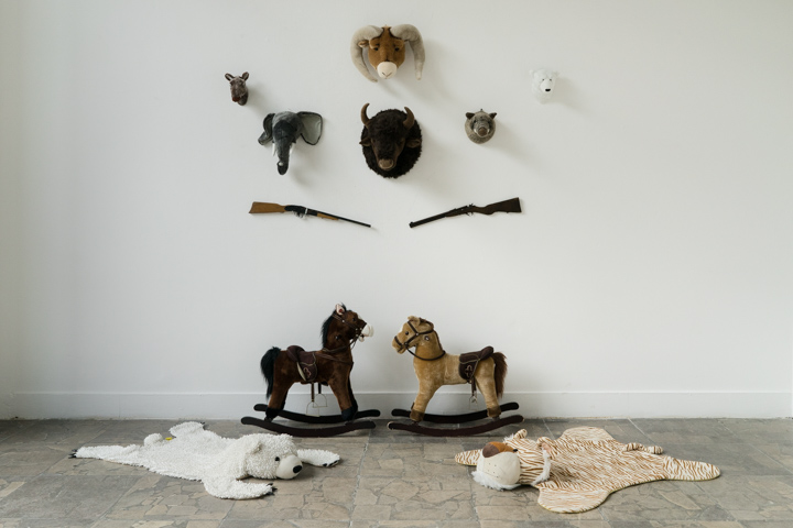 Zdjęcie przedstawia instalację składającą się z zabawek: pluszowych poroży zwierząt, strzelb. Na podłodze stoją dwa koniki na biegunach a przed nimi leżą pluszowe "niedźwiedzie" skóry. 