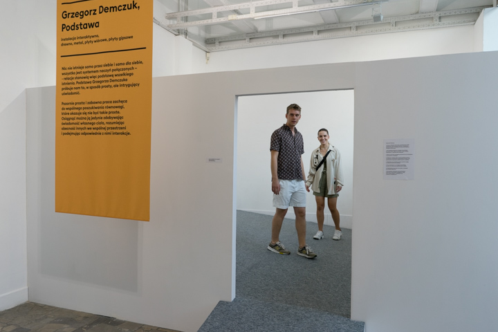 Zdjęcie przedstawia pracę Grzegorza Demczuka Podstawa. Widzimy zabudowaną białą przestrzeń, wewnątrz której znajduje się ruchoma podłoga, pokryta szarą wykładziną. W głębi widzimy dwoje ludzi: artystę Grzegorza Demczuka i młodą kobietę. Po lewej stronie jest zawieszona żółta plansza z opisem pracy. 
