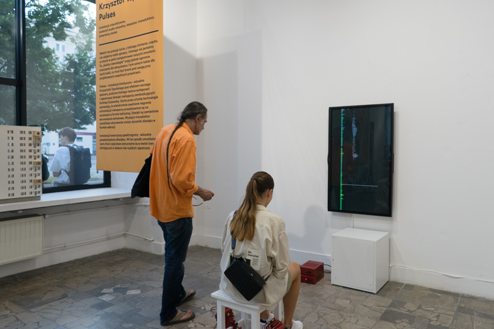 Zdjęcie przedstawia fragment ekspozycji z pracą Krzysztofa Topolskiego Pulses . Widzimy dwoje ludzi: stojącego mężczyznę w pomarańczowej koszuli i siedzącą na białym, drgającym taborecie dziewczynę w jasnym płaszczu. Oboje patrzą na ekran zawieszonego w pionie telewizora, na którym widać spektrogramy – wizualne przedstawienia dźwięku. Po lewej stronie jest zawieszona żółta plansza z opisem pracy. 