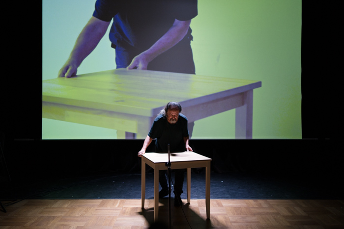 Na zdjęciu widać ciemną salę. Na środku stoi artysta, ręce opiera na drewnianym stole. Za nim znajduje się ekran, na którym widać w powiększeniu ten sam stół i ręce artysty.