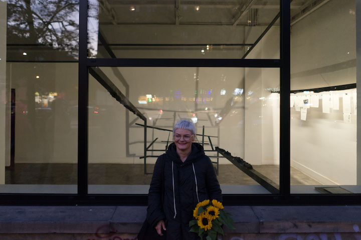 Zdjęcie wykonane wieczorem, na zewnątrz galerii.  Na tle instalacji widocznej przez okno widzimy ubraną na czarno autorkę wystawy trzymającą w ręku bukiet żółtych słoneczników. 