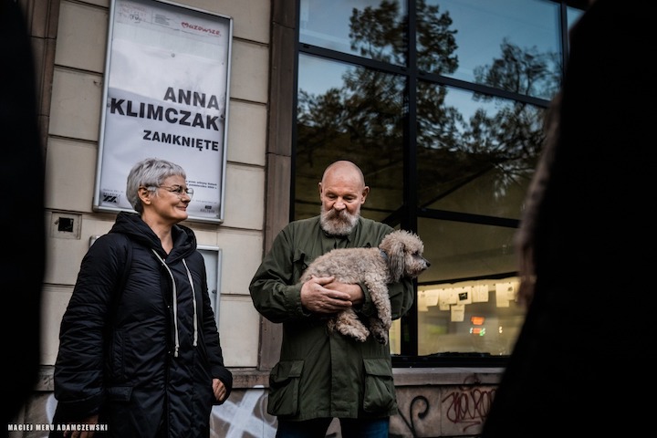 Na zdjęciu widzimy stojącą przed galerią Annę Klimczak i kuratora galerii Ryszarda Ługowskiego z kudłatym pieskiem na ręku. Za nimi widać fragment okna galerii i instalację artystki.