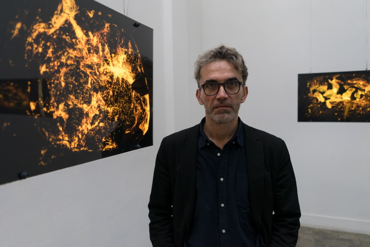 Na zdjęciu widzimy autora wystawy, stojącego na tle swoich prac. Z tyłu znajdują się dwie gabloty z podświetlonymi zdjęciami przedstawiającymi abstrakcyjne organiczne formy w żółciach, brązach i czerni. 
