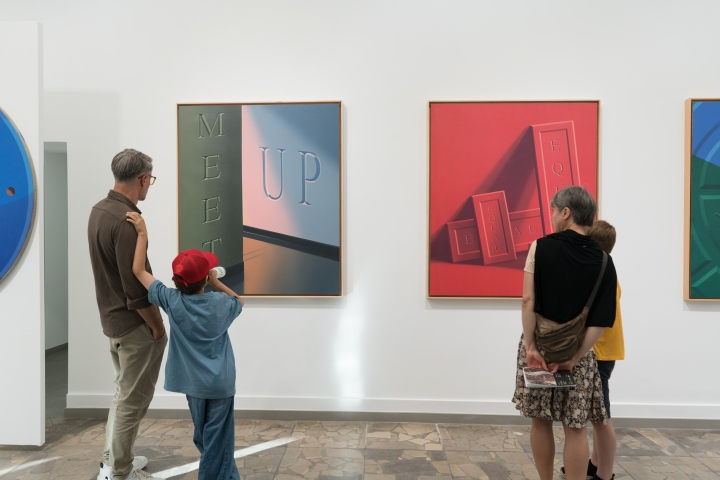Wnętrze galerii. Na ścianie galerii są powieszone dwa obrazy: jeden w kolorze szaro-niebieskim z napisem Meet up i drugi cały w czerwieniach z kilkoma napisami Equal. Przed obrazami po lewej i prawej stronie znajdują się widzowie.